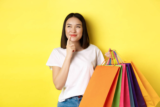 Giovane donna pensierosa sorridente incuriosita, mostrando borse della spesa, pensando di comprare qualcosa, in piedi su sfondo giallo - Foto, immagini