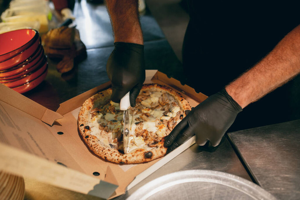 Zbliżenie. Szef kuchni krojący pyszną pizzę na kawałki w kartonowym pudełku. Wygląda smacznie, aromatycznie - Zdjęcie, obraz