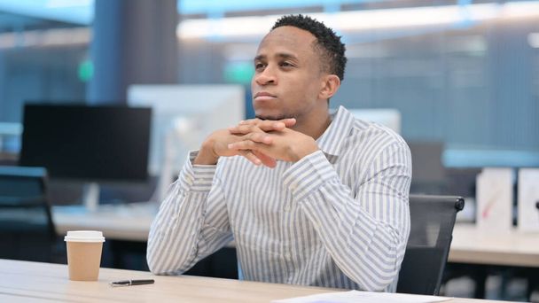 若いアフリカのビジネスマンがオフィスで考えて座っている - 写真・画像