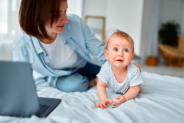 Stay at home, mom works remotely on laptop, taking care of her baby. A young mother on maternity leave tries to work in bed with a small child. Close up, copy space, background. - Photo, Image