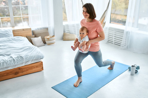 Eine sportliche Mutter beschäftigt sich mit dem Kind zu Hause mit Fitness und Yoga. Das Konzept von Sport, Mutterschaft und einem aktiven Lebensstil. Junge Frau im Sporttraining mit ihrem Kind. - Foto, Bild