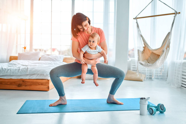A sports mother is engaged with the child in fitness and yoga at home. The concept of sports, motherhood and an active lifestyle. Young woman in sports training with her child. - Фото, изображение