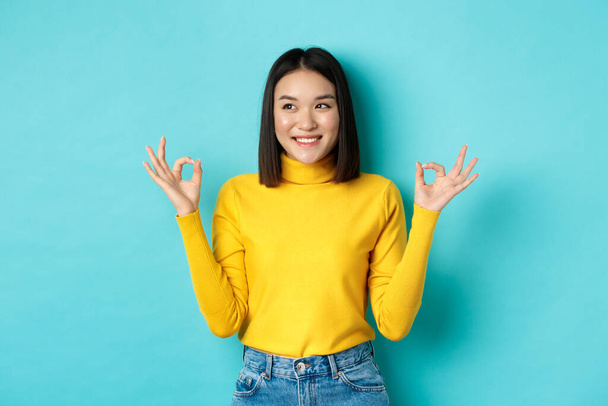 Piękna japońska dziewczyna w żółtym swetrze, pokazując Ok znaki i uśmiech, patrząc w lewo i logo i polecając firmę, stojąc na niebieskim tle - Zdjęcie, obraz