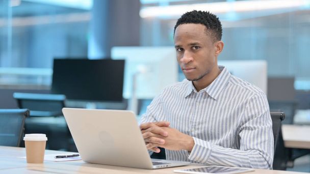 Attractive African Businessman Looking at Camera while using Laptop in Office - Photo, Image
