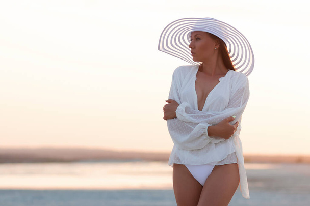 Sensual modelo joven con bikini blanco con camisa y sombrero elegante de pie en las luces del atardecer en la playa - Foto, imagen