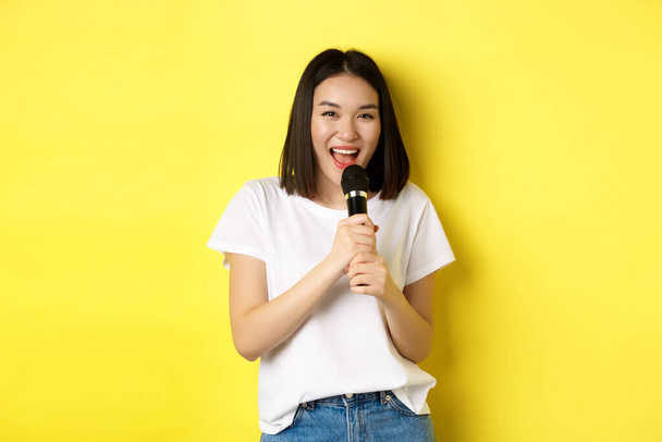 Красивая азиатка поет караоке, выступает с микрофоном, улыбается счастливой и смотрит в камеру, стоя на желтом фоне - Фото, изображение