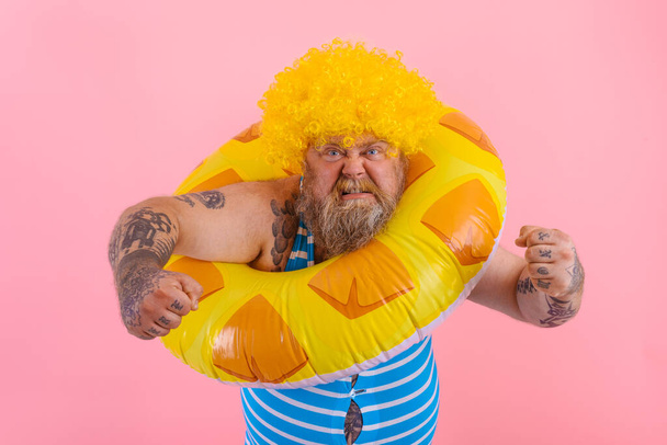 Gordo hombre enojado con peluca en la cabeza está listo para nadar con un donut salvavidas - Foto, imagen