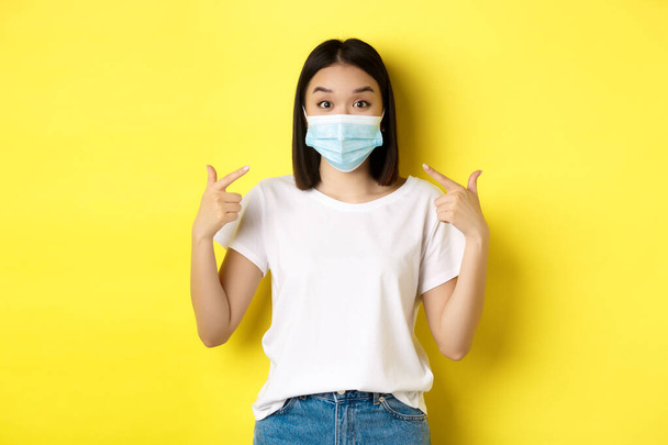 Covid-19 、隔離と社会的距離の概念。白いTシャツの若いアジア人女性,彼女の医療マスクを指しているコロナウイルスから,黄色の背景 - 写真・画像
