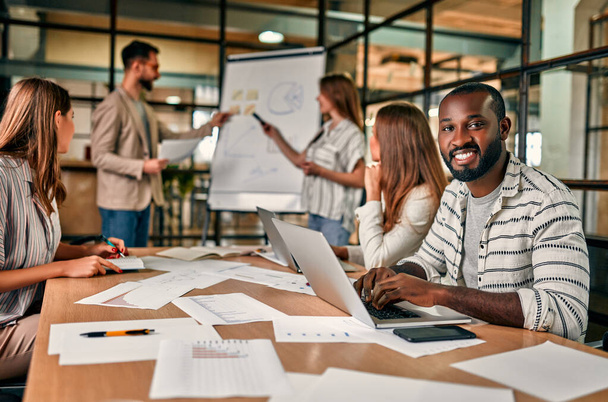 Młoda grupa ludzi omawia nowy biznes plan na tablicy, pracując na laptopach, siedząc w nowoczesnym biurze. Afroamerykanin pracuje ze swoimi kolegami w biurze.. - Zdjęcie, obraz
