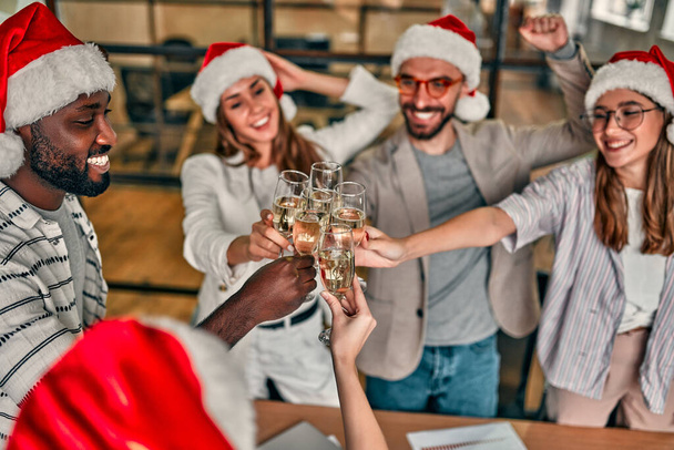 Hyvää joulua ja onnellista uutta vuotta! Monirotuiset nuoret luovat ihmiset juhlivat lomaa nykyaikaisessa toimistossa. Ryhmä nuoria liikemiehiä juo samppanjaa yhteistyössä. - Valokuva, kuva