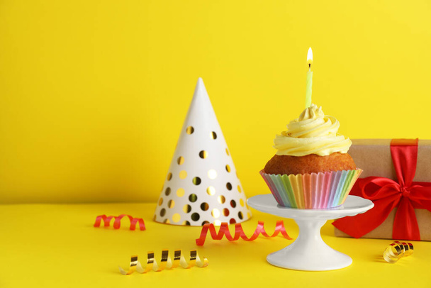 Delicious birthday cupcake with candle near gift box and party hat on yellow background, space for text - Foto, imagen