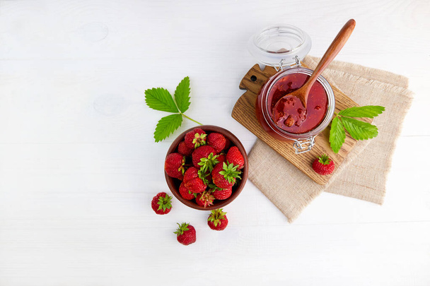 Strawberry jam in a glass jar and fresh berries in a ceramic bowl. Homemade strawberry marmalade. Conservation and summer harvesting. Top view. - Foto, Imagen