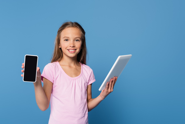 Kid holding digital tablet and showing smartphone isolated on blue  - 写真・画像