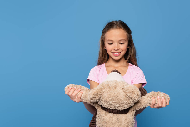 Positive kid looking at teddy bear isolated on blue  - Фото, изображение