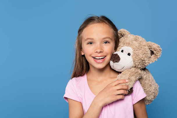 Kid with soft toy smiling at camera isolated on blue  - Fotografie, Obrázek