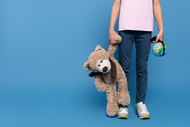 Cropped view of kid holding teddy bear and globe on blue background  - Foto, Imagen
