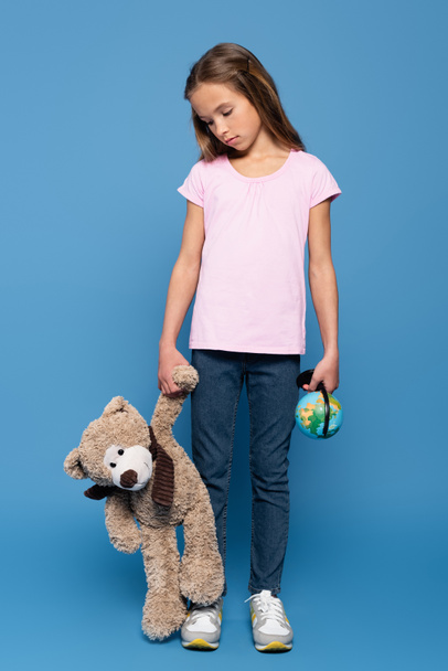Грустный ребенок с глобусом смотрит на плюшевого мишку на синем фоне  - Фото, изображение