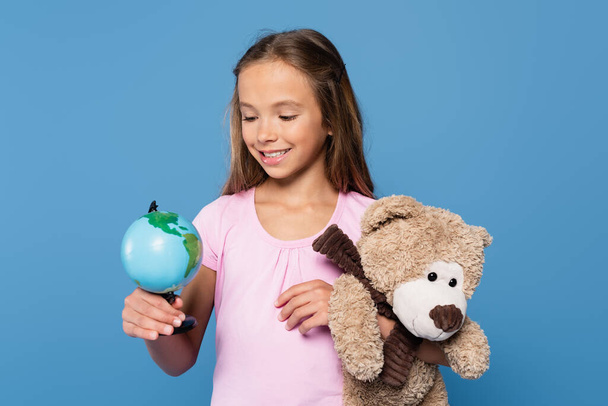 Hübsches Mädchen mit Globus und Teddybär lächelt isoliert auf blau - Foto, Bild