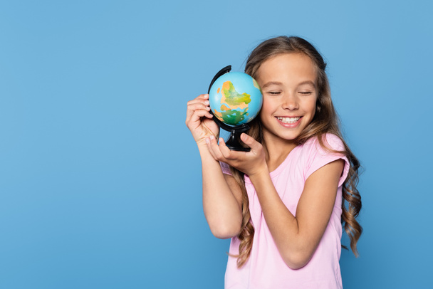 Girl with closed eyes smiling while holding globe isolated on blue  - Foto, imagen