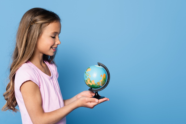 Zijaanzicht van het lachende meisje dat de wereldbol vasthoudt aan handen geïsoleerd op blauw  - Foto, afbeelding