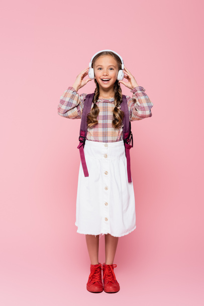 Positive schoolkid holding wireless headphones on pink background  - Foto, afbeelding
