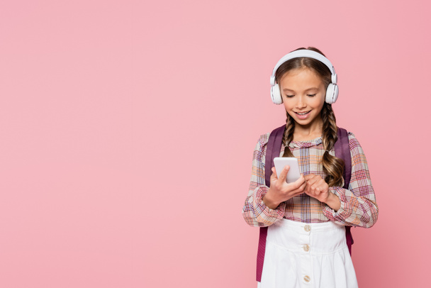 Smiling schoolchild in headphones using cellphone isolated on pink  - Photo, Image