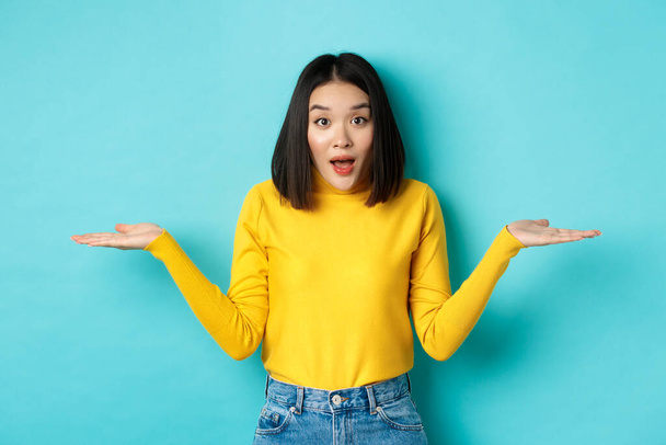Image of indecisive asian woman shrugging shoulders, spread hands sideways and looking clueless at camera, standing confused against blue background - 写真・画像