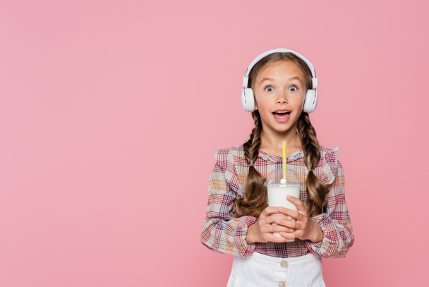 Criança excitada em fones de ouvido segurando milkshake isolado em rosa  - Foto, Imagem