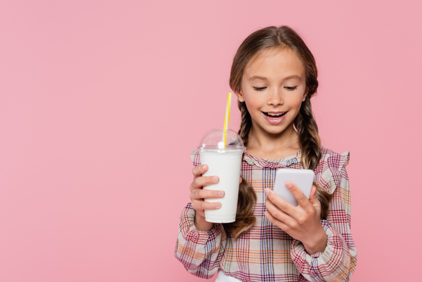 Mosolygós gyerek használ okostelefon és gazdaság tejturmix izolált rózsaszín  - Fotó, kép