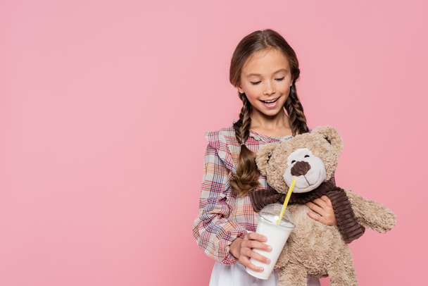Kid in plaid shirt holding milkshake near teddy bear isolated on pink  - Foto, Imagen