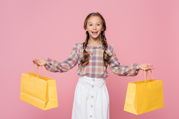 Positive kid holding yellow shopping bags isolated on pink  - Φωτογραφία, εικόνα