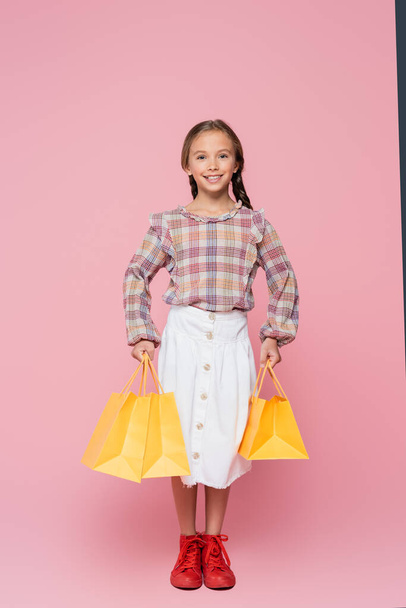 visão de comprimento total da menina pré-adolescente feliz com sacos de compras amarelos no fundo rosa - Foto, Imagem