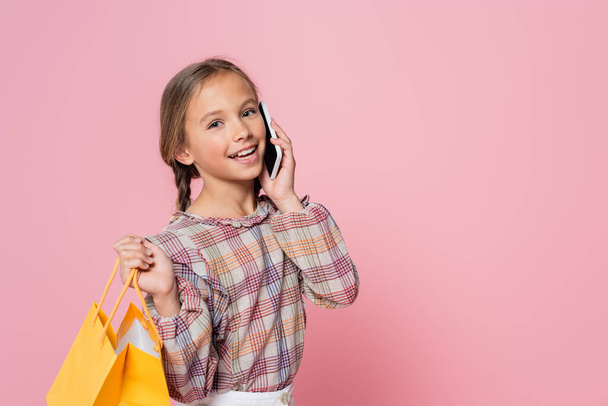 preteen girl with yellow shopping bag talking on mobile phone isolated on pink - 写真・画像