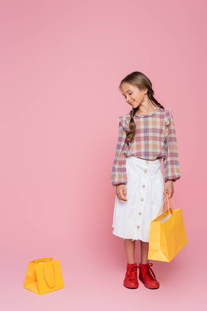 smiling girl in white skirt and plaid blouse looking at yellow shopping bag on pink background - Photo, Image