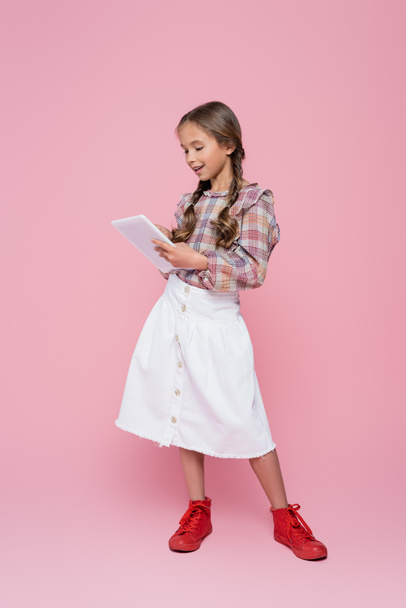 preteen child in white skirt and plaid blouse using digital tablet on pink background - Фото, изображение