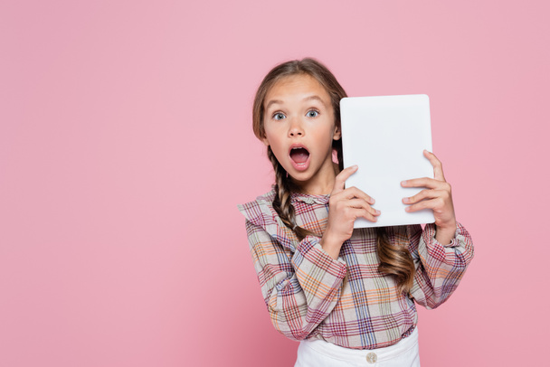 amazed girl looking at camera while holding digital tablet isolated on pink - Фото, изображение