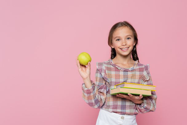 pleased girl with green apple and textbooks smiling at camera isolated on pink - Photo, image