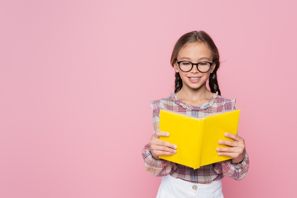 smiling girl in eyeglasses and plaid blouse reading book isolated on pink - Foto, Imagen