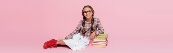 χαρούμενο κορίτσι με καρό μπλούζα και λευκή φούστα κάθεται σε ροζ φόντο κοντά σε στοίβα από βιβλία, πανό - Φωτογραφία, εικόνα