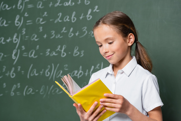 pleased schoolkid reading book near blurred equations on chalkboard - Foto, Imagen
