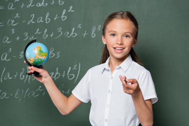 счастливый школьник с маленьким глобусом, указывая карандашом на камеру рядом с доской с уравнениями - Фото, изображение