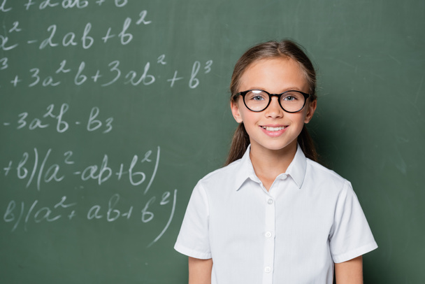 smiling schoolgirl in eyeglasses looking at camera near chalkboard with equations - Photo, image