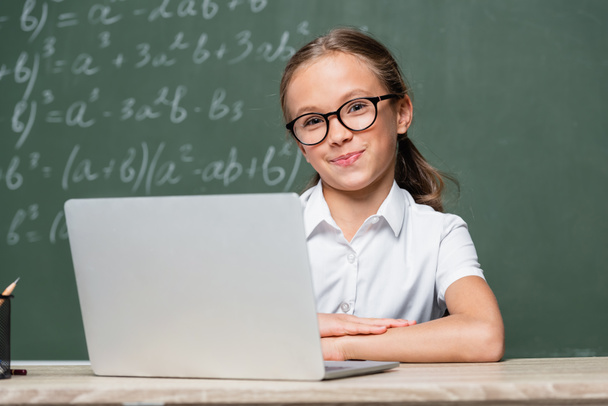schoolgirl in eyeglasses smiling at camera near laptop and chalkboard on blurred background - Photo, image