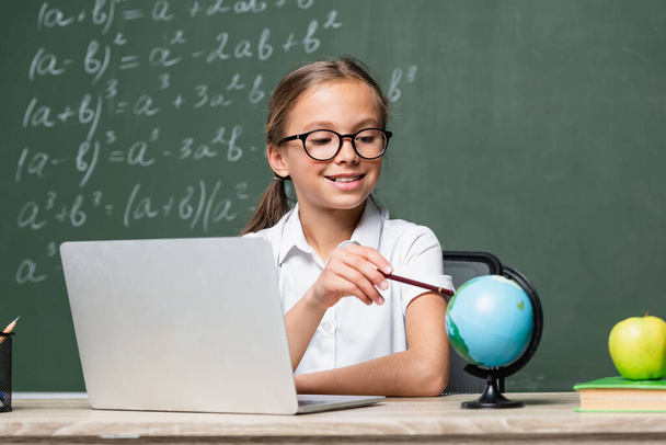 smiling schoolkid pointing with pencil at globe near laptop and blurred chalkboard - Foto, Bild