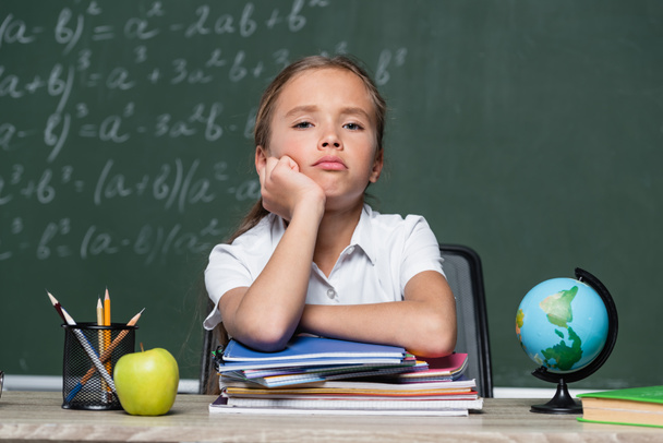 upset schoolgirl looking at camera near notebooks, globe and chalkboard on blurred background - Foto, imagen
