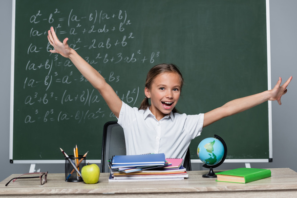 cheerful schoolgirl with open arms near notebooks on desk and chalkboard with equations - Foto, Imagen