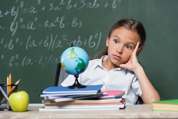 thoughtful schoolgirl looking at globe near notebooks and chalkboard on blurred background - Photo, Image