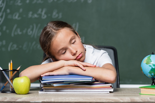 κουρασμένη μαθήτρια κοιμάται σε σημειωματάρια κοντά στον κόσμο και μήλο στο γραφείο - Φωτογραφία, εικόνα