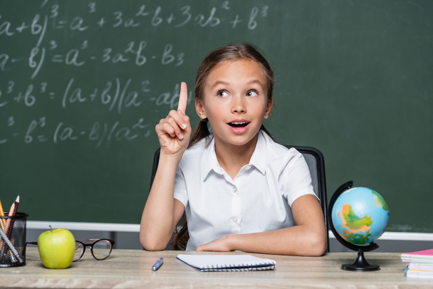 amazed schoolgirl showing idea gesture near globe, notebook and blurred chalkboard - Photo, image