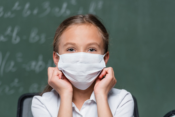 preteen schoolgirl in protective mask looking at camera near blurred chalkboard on background - Photo, image
